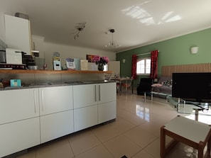 A modern kitchen with white cabinets, a dining area with a table and chairs, a television, and a green wall. - Entire private gîte (Lédenon)