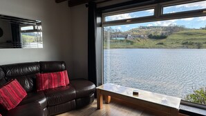 A living room with a leather sofa, a wooden coffee table, and a large window overlooking a lake and hills. - Wallace Lodge - Unique experience (Comrie)