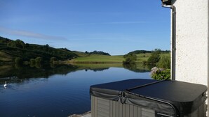 A hot tub on a patio overlooking a lake with a swan, surrounded by greenery and hills. - Wallace Lodge - Unique experience (Comrie)