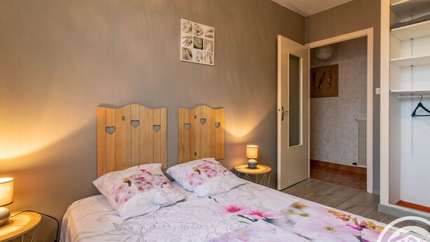 Ein Schlafzimmer mit Bett, Holz-Kopfteil mit Herzformen und einer Nachttischlampe sowie einer blumigen Tagesdecke.