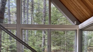 A modern interior with a wooden floor, a staircase with circular cutouts, and large windows offering a forest view. - A Prism in the Wood (Mount Desert)