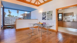 A modern kitchen with a built-in oven, a dining area with a glass table and metal chairs, and a view of the outdoors through a large window.