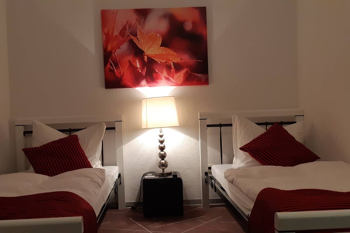 Ein Hotelzimmer mit zwei Einzelbetten, jedes mit einer rot-weiß gestreiften Decke, einem roten Kopfkissen und einem weißen Kopfkissen. Zwischen den Betten befindet sich eine Wandlampe, darüber ein Bild mit Herbstlaub.