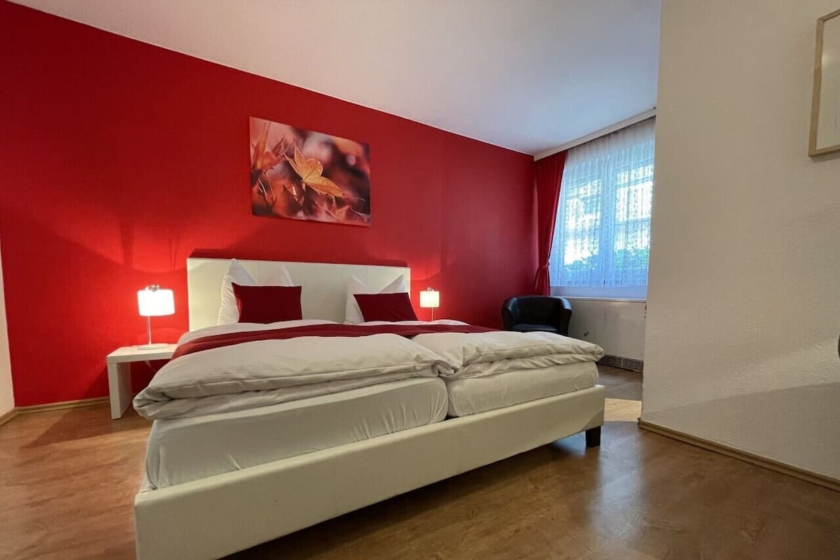 Ein Schlafzimmer mit einem Bett, zwei Nachttischen, einem Fenster mit Vorhängen und einem Bild an der Wand. Eine rote Wand ist ebenfalls zu sehen.