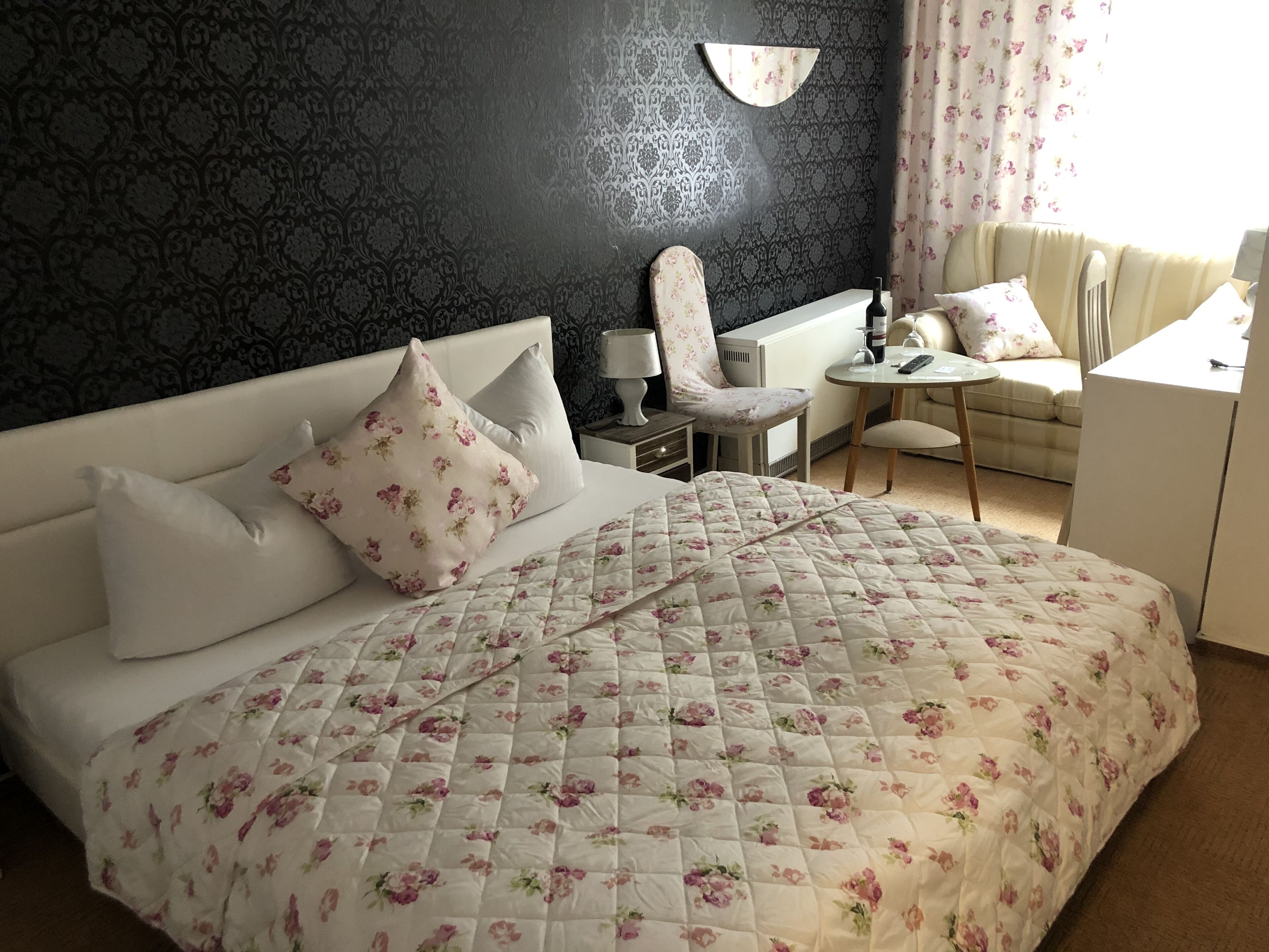 A neatly made bed with floral bedding, a bedside table with a lamp, a chair, a small table with a remote control, and a sofa.