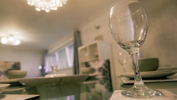 A dining area with a clear glass wine glass on a table, a chandelier overhead, and a blurred background with shelves and a window. - Stunning Holiday Stay With Free Gym and Parking (Cardiff)