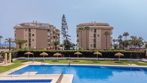 A swimming pool with a metal fence, surrounded by palm trees and a building with multiple balconies. - Beach front apartment in Torrox Costa with WiFi (Torrox Costa)