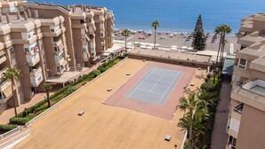 A tennis court surrounded by residential buildings with balconies, overlooking a beach with umbrellas and palm trees. - Beach front apartment in Torrox Costa with WiFi (Torrox Costa)