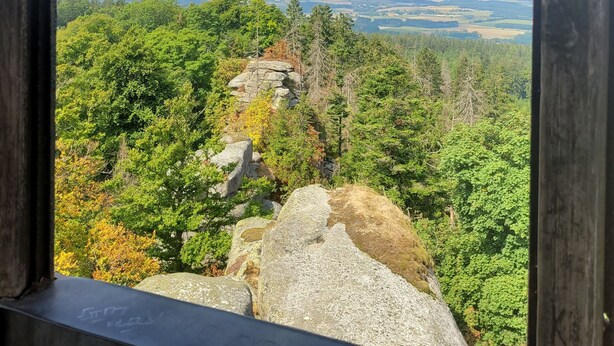 Une vue à travers une structure en bois ornée d’une arche gothique, donnant sur un paysage boisé où l’on aperçoit des formations rocheuses et des champs éloignés.