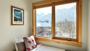 Ein Schlafzimmer mit einem großen Fenster, das Blick auf schneebedeckte Berge und eine Holzhütte bietet. An der Wand hängt ein gerahmter Poster von Lake Louise.