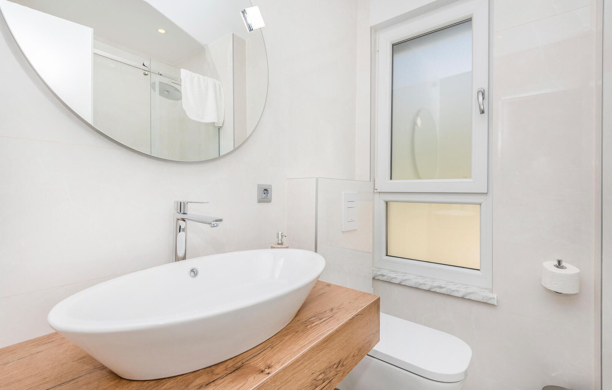 Une salle de bain moderne avec un lavabo blanc sur pied, un meuble-lavabo en bois, des toilettes, un grand miroir et une fenêtre en verre dépoli.