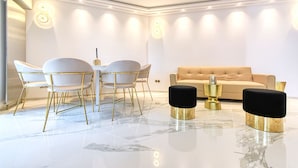 Una sala de estar moderna con un sofá color beige, sillas blancas con detalles dorados, piso de mármol y una mesa de centro de vidrio.