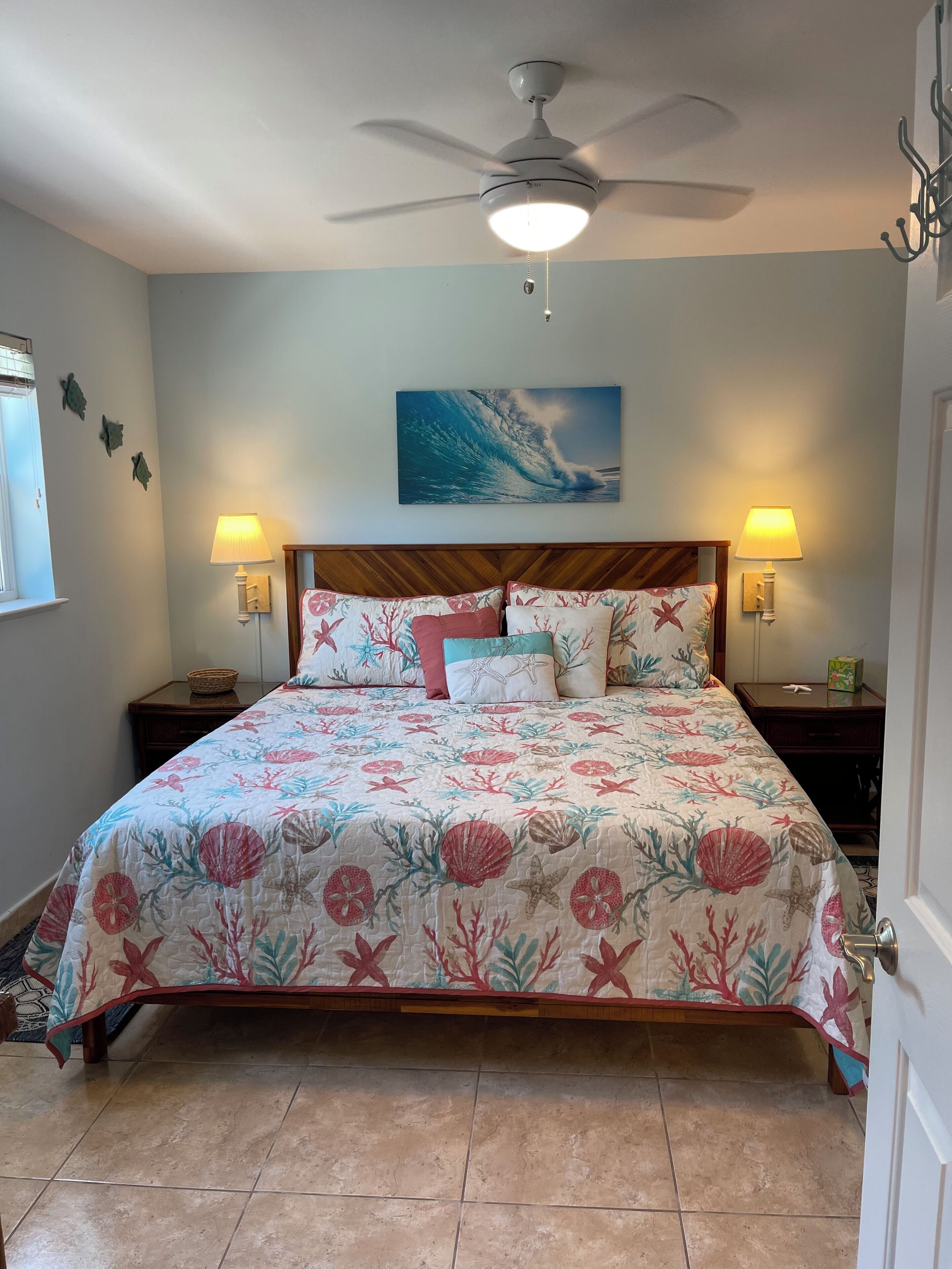 A bedroom with a bed, two bedside tables, a ceiling fan, a wall-mounted painting, and a window.