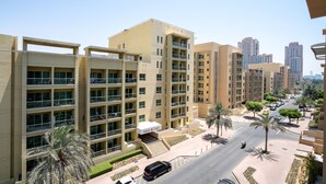 Un edificio residencial de varios pisos con balcones, una entrada central y una calle con coches y palmeras.