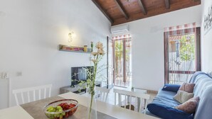 Una sala de estar moderna con un sofá, una mesa de comedor con frutero, un televisor y un balcón con vistas.