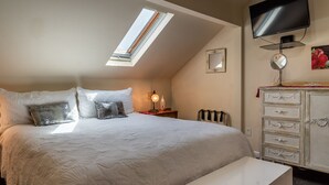 Une chambre avec un plafond en pente, un lit avec une literie blanche, une table de chevet avec une lampe, une commode, un téléviseur et un puits de lumière.