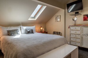 A bedroom with a sloped ceiling, a bed with white bedding, a bedside table with a lamp, a dresser, a television, and a skylight.