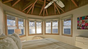 Une chambre dotée d’un grand lit, d’un ventilateur de plafond et de plusieurs fenêtres donnant sur l’océan.