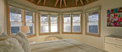 A bedroom with a large bed, a ceiling fan, and multiple windows offering ocean views.