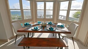Un coin repas avec vue sur la mer, comprenant une table en bois dressée pour quatre personnes, avec des serviettes et des assiettes bleues, des chaises blanches et un banc.