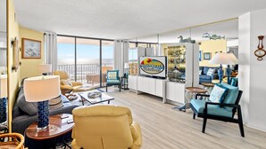 Un salon spacieux avec une grande fenêtre donnant sur l’océan, une enseigne indiquant « North Beach Vacations », et un coin salon confortable avec un fauteuil jaune et un canapé bleu.