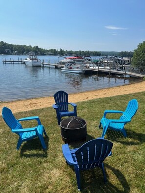 A lakeside dock with several boats, a grassy area with blue Adirondack chairs, and a round table. - Winnisquam Lakeside Funky Ski Chalet w-Sandy Beach & Boat Docks (Tilton)