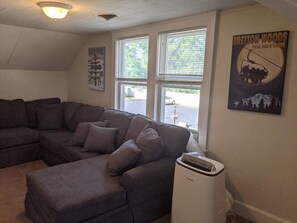 A living room with a sectional sofa, a ceiling light, a window with blinds, and a poster on the wall. - Winnisquam Lakeside Funky Ski Chalet w-Sandy Beach & Boat Docks (Tilton)