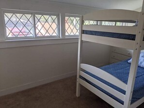 Ein Etagenbett mit unterem Bett und einer Kommode darunter. Drei Fenster mit Metallgitterstäben sind vorhanden.