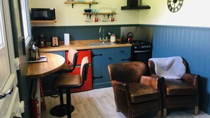 Une cuisine compacte équipée d’une cuisinière, d’un four à micro-ondes et d’étagères en bois. On y trouve deux fauteuils en cuir et une petite table.