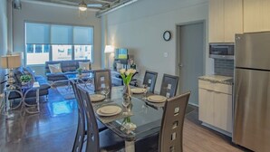 A modern kitchen with stainless steel appliances, a dining area with a glass table, and a living space with a sofa and TV.
