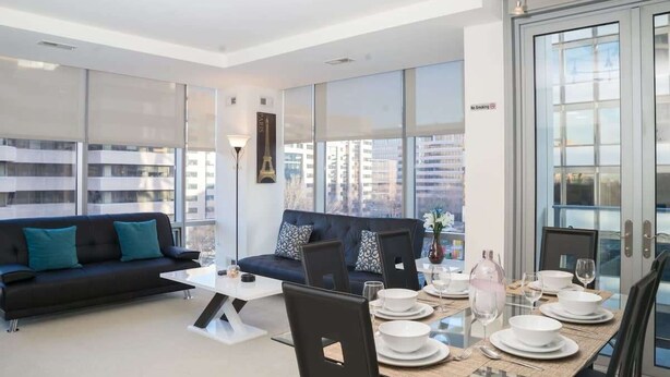 Een moderne woonkamer met een bank, eettafel en stoelen. Er zijn grote ramen met uitzicht op gebouwen.