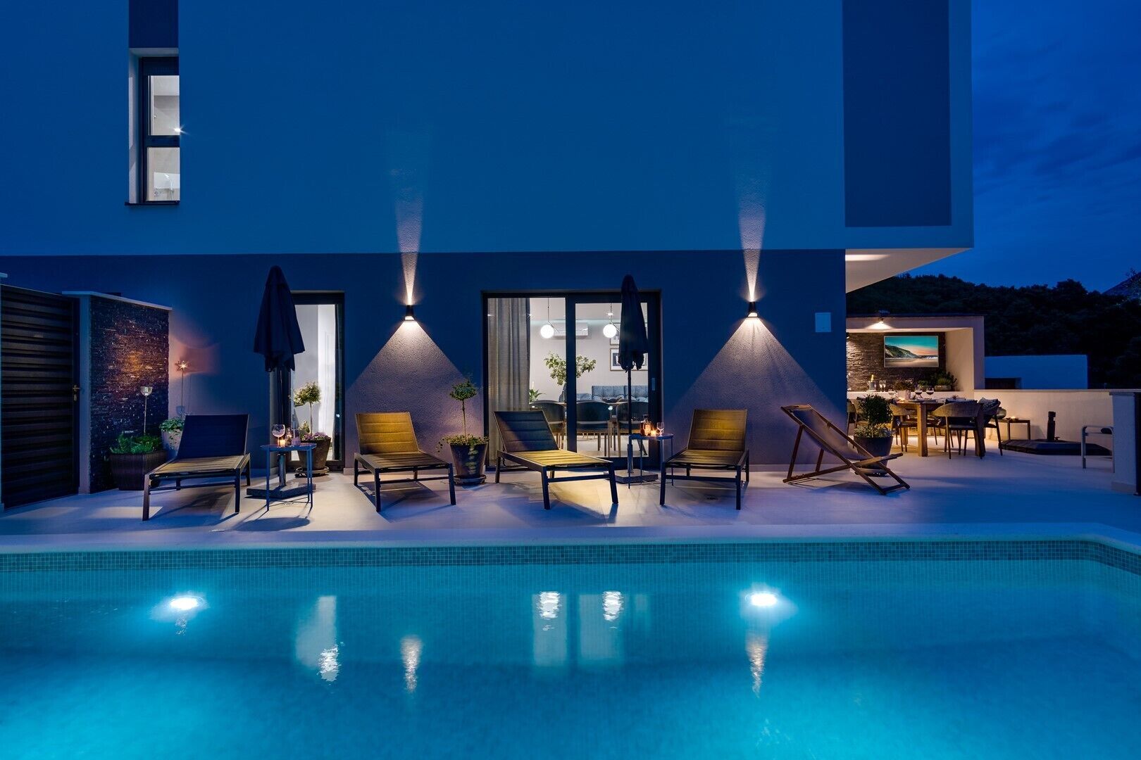 A modern outdoor pool area with lounge chairs and a dining table, surrounded by a blue building with large windows and a view of the landscape.