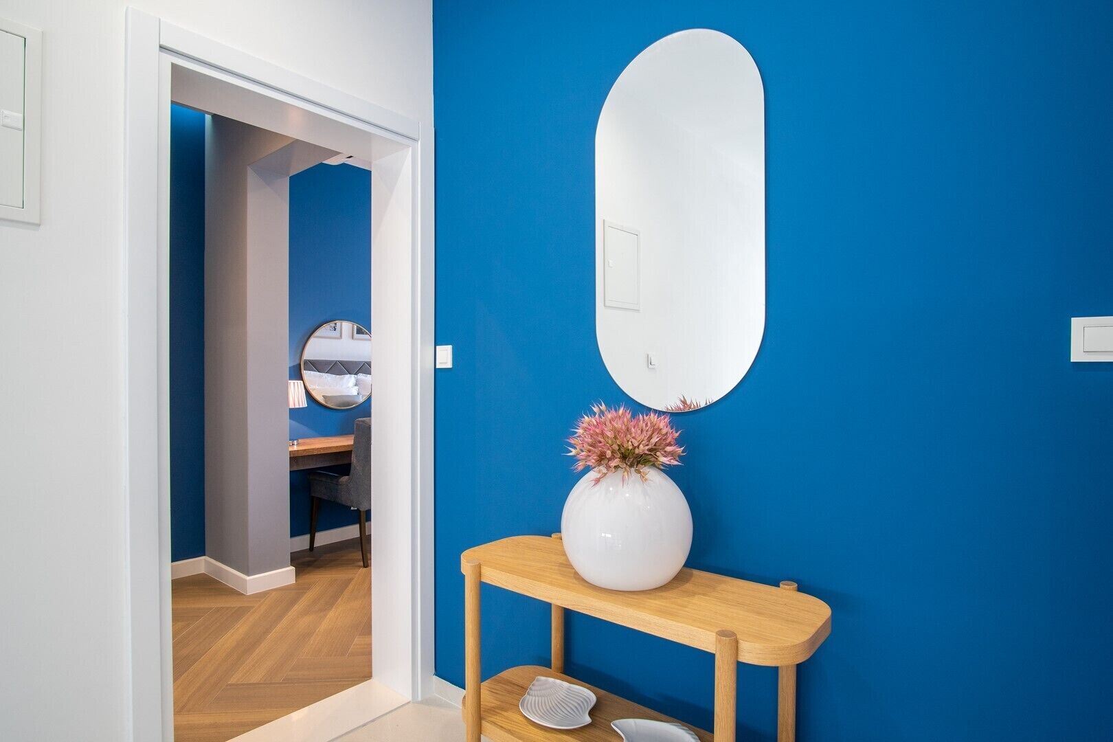 Een kamer met een blauwe wand, een ronde witte spiegel, een houten console tafel met een bloemenvaas, en een bed met een grijze hoofdbord.