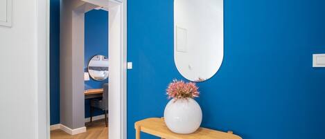 Een kamer met een blauwe wand, een ronde witte spiegel, een houten console tafel met een bloemenvaas, en een bed met een grijze hoofdbord.