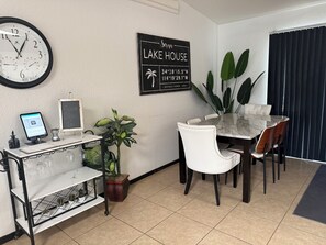 A hotel room with a dining table, chairs, a clock, a sign indicating the hotel's name and location, a computer on a desk, and a potted plant.