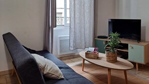Een woonkamer met een donkere bank, een houten salontafel, een televisie op een standaard en een raam met gordijnen.