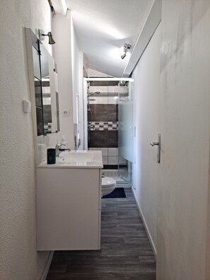 Et aflangt badeværelse med en hvid kommode, et glasbadeværelsesafskærmning og et synligt toilet.