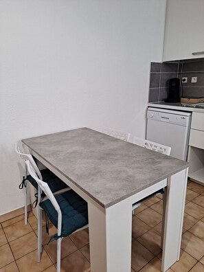Et lille køkken med et hvidt bord og stole, kaffemaskine og opvaskemaskine.