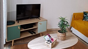 Een woonkamer met een televisie op een houten standaard, een bank met een mosterdkleurig kussen, een ronde salontafel met een kaart houder, en een potplant.