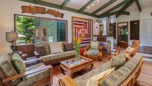 A spacious living room with wooden furniture, a large window, and a decorative wall hanging. - Hanalei Happy Place, Incomparable Home TVNC#1158 (Hanalei)