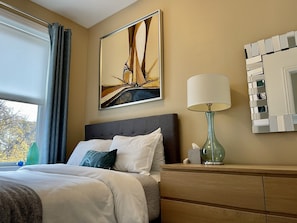 Habitación de hotel con una cama, una mesita de noche con lámpara, un cuadro en la pared y una ventana con cortinas.