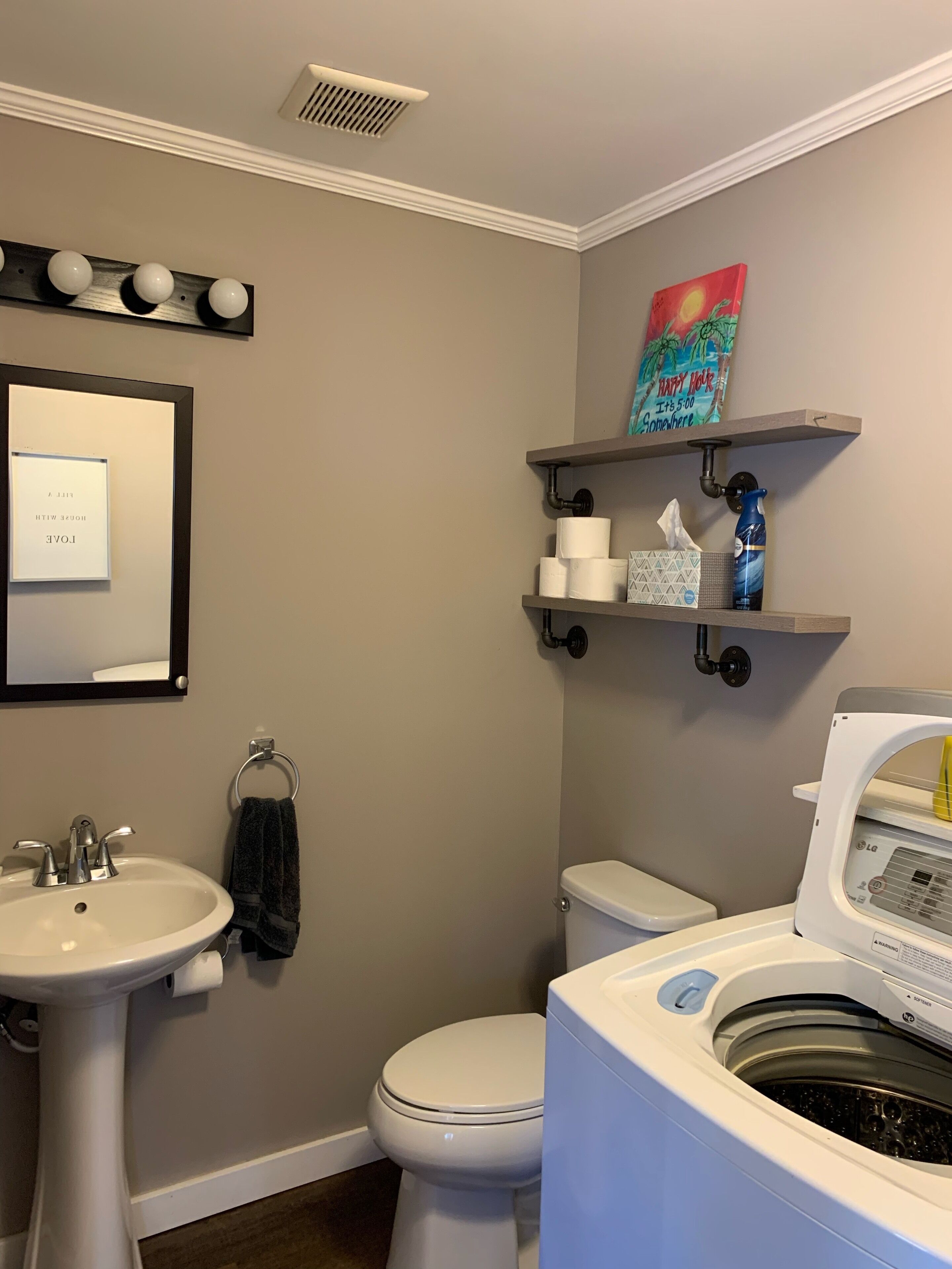 A bathroom with a sink, toilet, and washing machine. There is a towel rack and a shelf with toilet paper and a bottle.