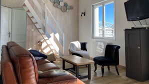 Un salon avec un escalier en bois, un canapé, un fauteuil, une table et un téléviseur posé sur un meuble.