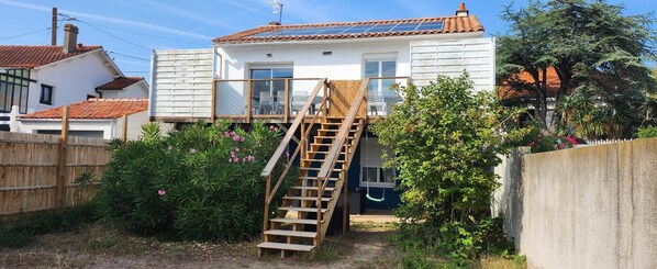 Une maison avec un escalier en bois menant à une terrasse, entourée de verdure et bordée d’un mur en béton.