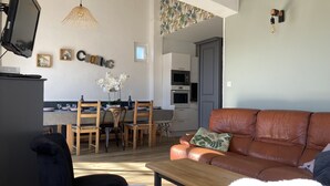 Un salon moderne avec un canapé en cuir marron, un fauteuil bleu et une table basse en bois. On aperçoit à l’arrière-plan un coin repas avec une table et des chaises en bois.