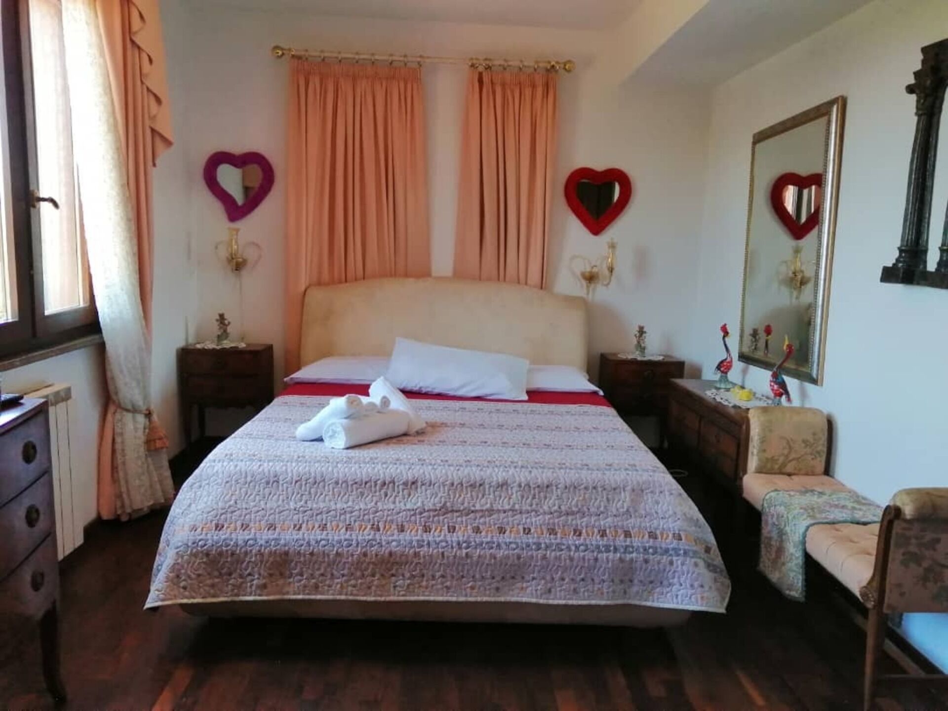 Un letto rifatto con una coperta a motivi, due cuscini e un asciugamano piegato. Sono presenti due comodini con lampade, una cassettiera e una sedia. La stanza ha tende e decorazioni a forma di cuore.
