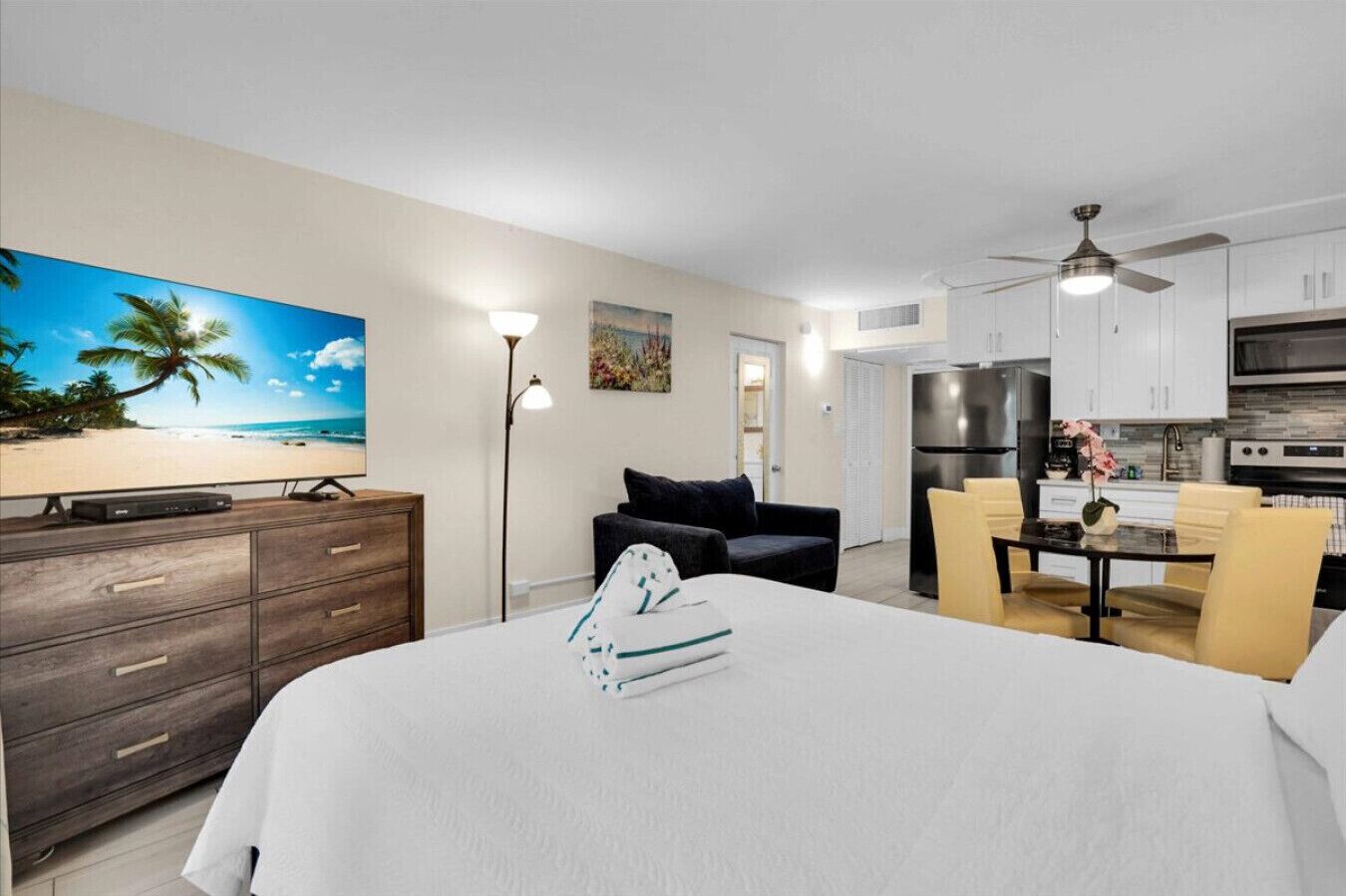 A hotel room with a bed, a desk, a chair, a dining table with chairs, a ceiling fan, and a kitchen area with cabinets and appliances.