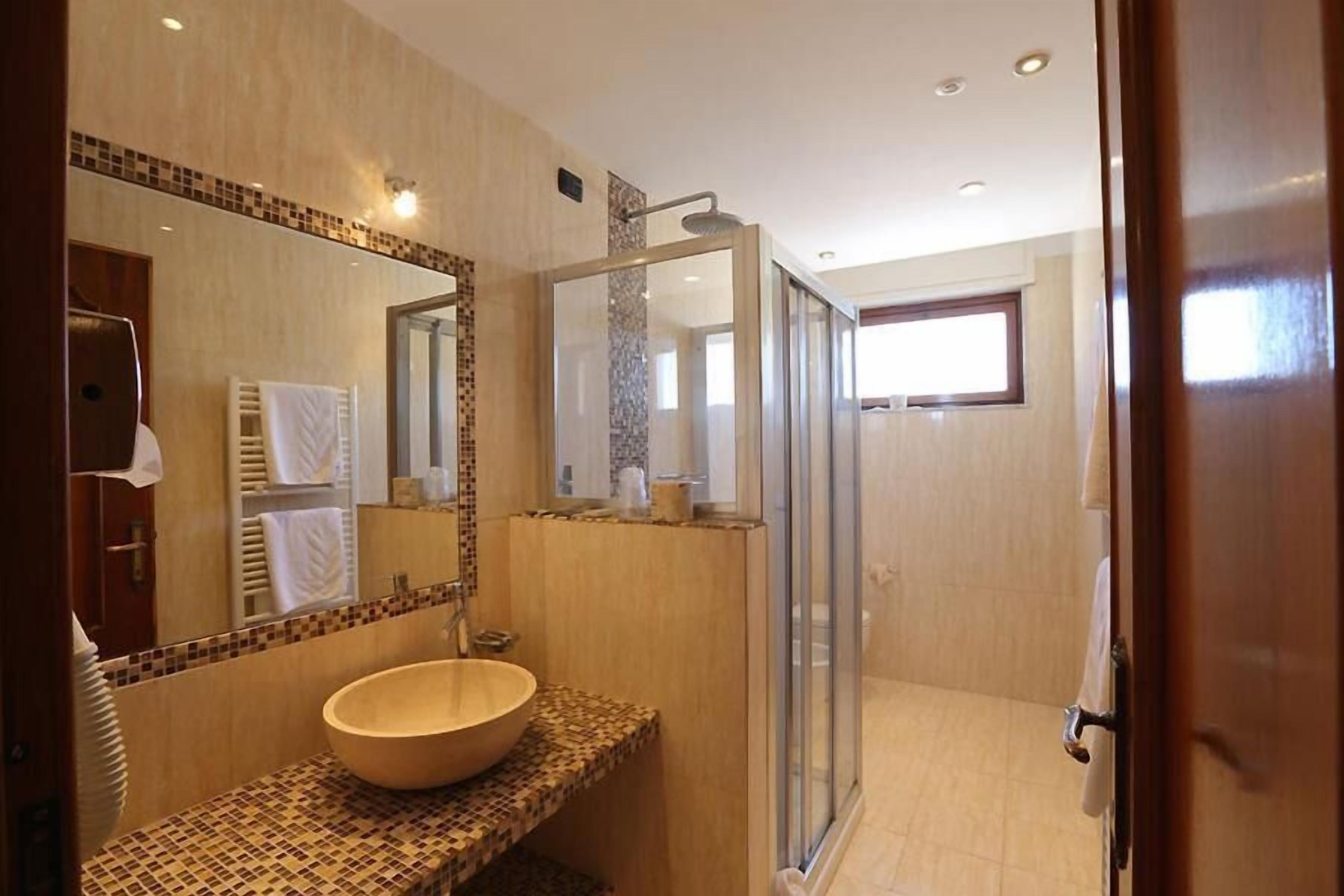 A modern bathroom with a large mirror, a wooden vanity, a glass shower enclosure, and a view of the adjacent room.