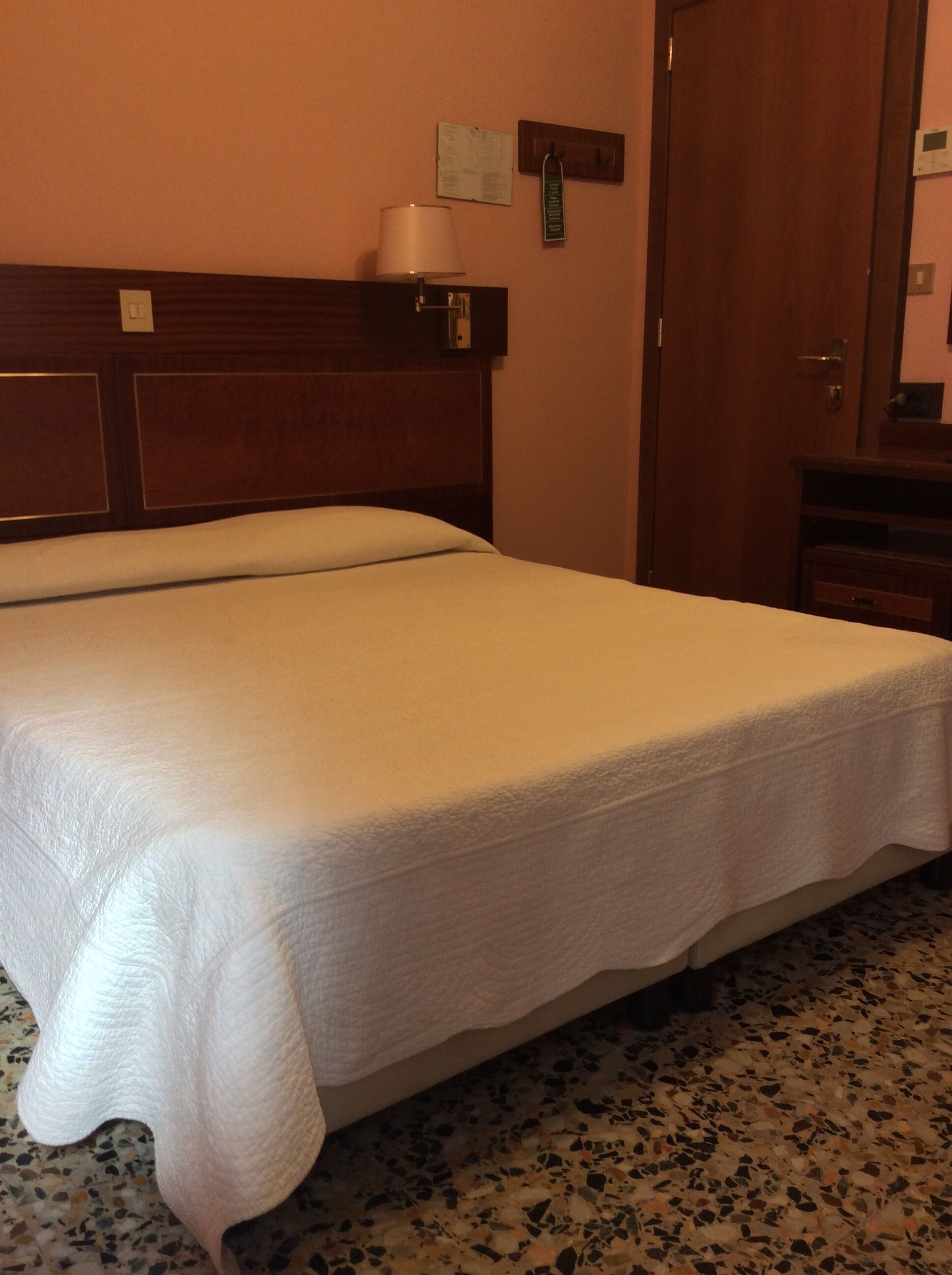 Una habitación de hotel con una cama grande, un cabecero de madera, una mesita con una lámpara y una puerta.