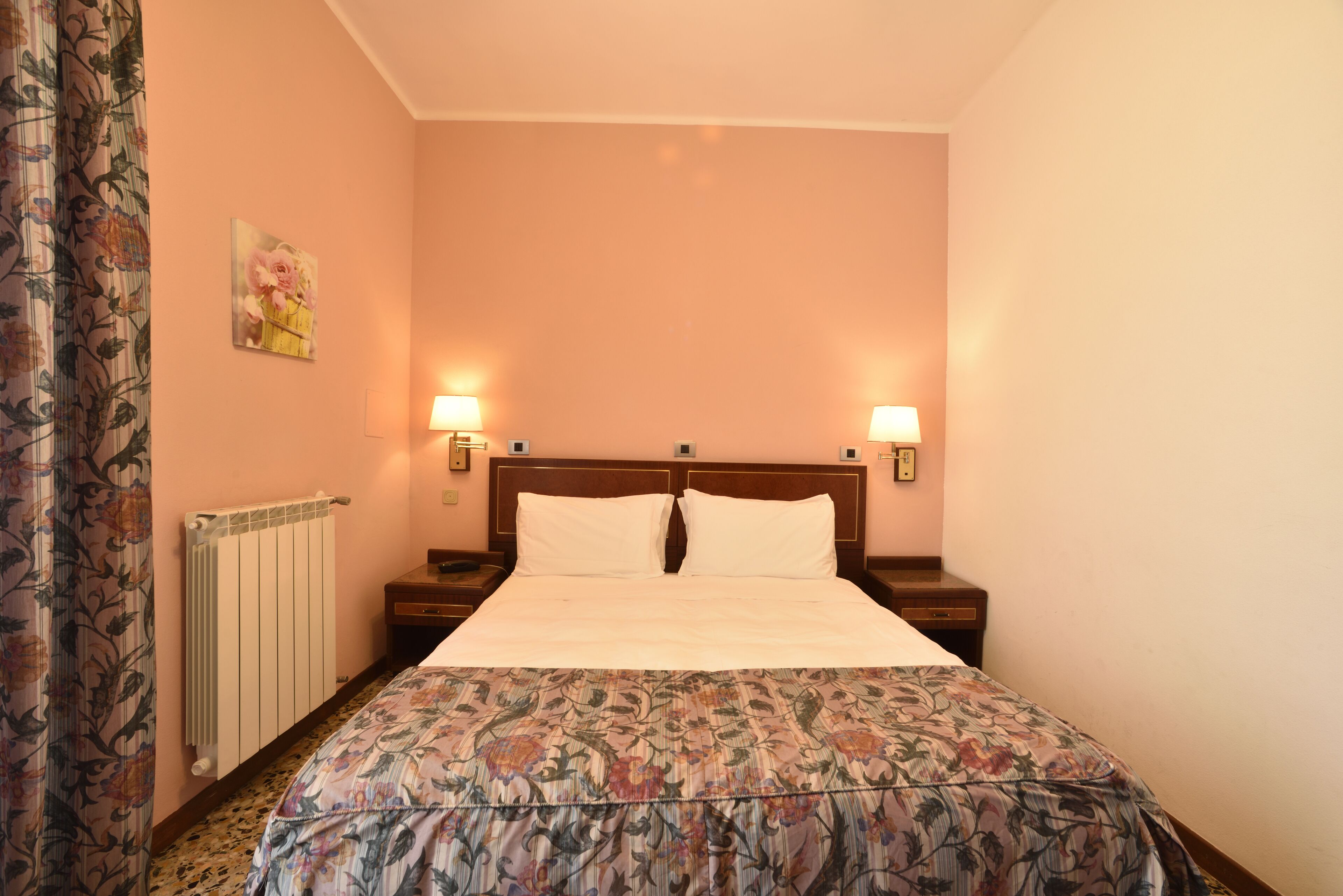Camera d'albergo con un letto grande, due comodini, un radiatore e una coperta con motivo floreale.